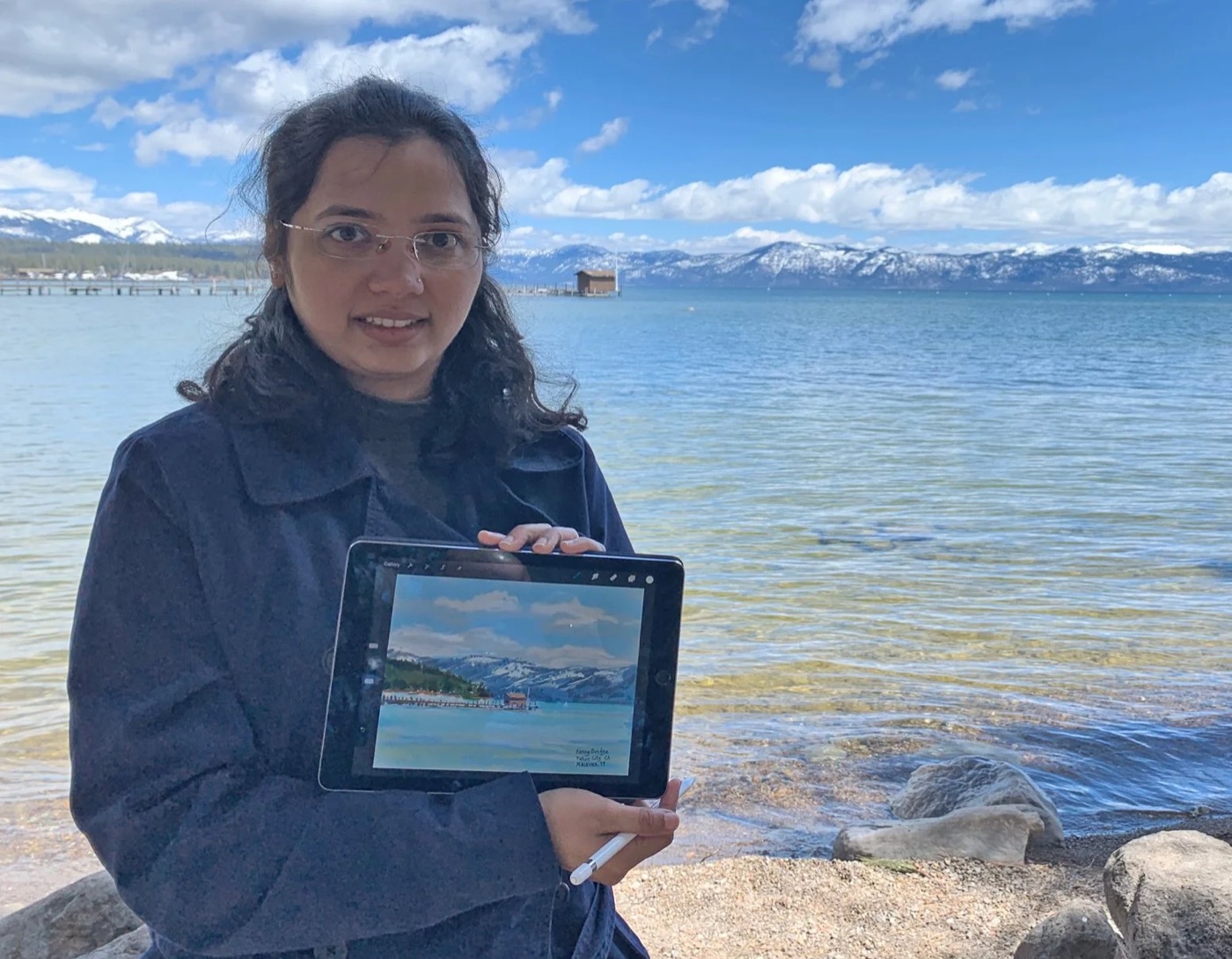 Malavika standing on the shore of Lake Tahoe, holding up an iPad showing the painting she has just made of the view behind her.